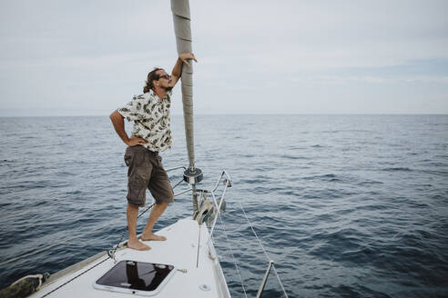 Männlicher Seemann steht auf dem Bug eines Segelbootes im Meer gegen den Himmel - GMLF00290