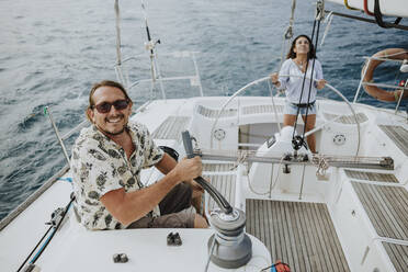 Man maneuvering with winch while woman driving sailboat in sea - GMLF00304