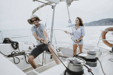 Man maneuvering with winch while woman driving sailboat against sky - GMLF00305