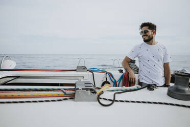 Smiling young man wearing sunglasses standing in sailboat against sky - GMLF00306