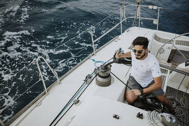 Young sailor wearing sunglasses maneuvering with winch in sailboat - GMLF00310