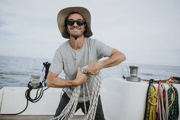 Smiling sailor wearing sunglasses and hat holding ropes in sailboat - GMLF00311