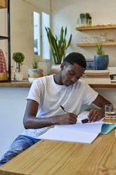 Young man writing notes in book on table while sitting at home - VEGF02400