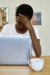 Young woman with head in hand using laptop on table at home - VEGF02412
