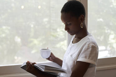 Nahaufnahme einer jungen Frau, die eine Kaffeetasse hält und zu Hause am Fenster ein Buch liest - VEGF02421