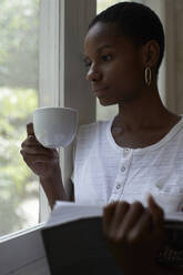 Nahaufnahme einer nachdenklichen jungen Frau, die einen Kaffee und ein Buch in der Hand hält, während sie durch das Fenster ihres Hauses schaut - VEGF02422