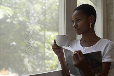 Nahaufnahme einer nachdenklichen Frau mit Kaffee und Buch, die durch das Fenster ihres Hauses schaut - VEGF02423