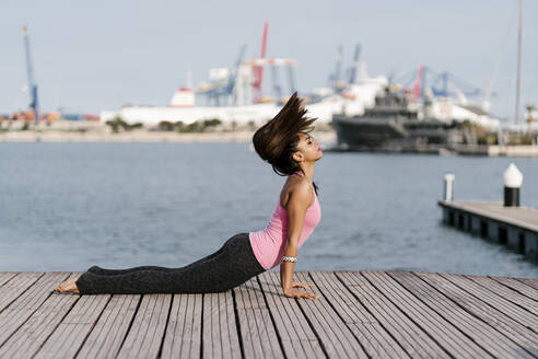 Female athlete with eyes closed practicing cobra pose on pier against sea at harbor - EGAF00318
