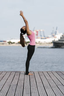 Female athlete practicing upward tree position while standing on pier against sea - EGAF00321