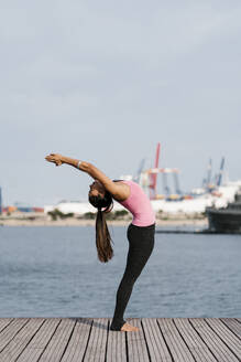 Female athlete practicing upward tree position while standing on pier against sea - EGAF00323