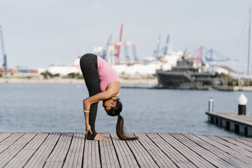 Female athlete practicing standing forward bend pose on pier against sea at harbor - EGAF00324