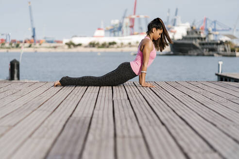 Female athlete practicing cobra pose on pier at harbor - EGAF00325