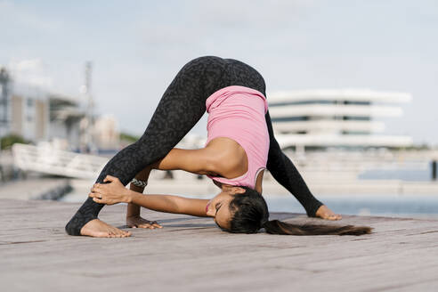 Female athlete practicing wide-legged forward bend on pier against sky - EGAF00326