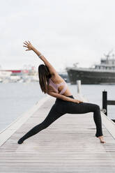 Female athlete with legs apart practicing yoga on pier at harbor - EGAF00332