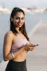 Nahaufnahme einer lächelnden Sportlerin, die ein Smartphone benutzt, während sie am Hafen steht - EGAF00335
