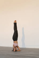Female athlete practicing headstand on hardwood floor against wall - EGAF00345