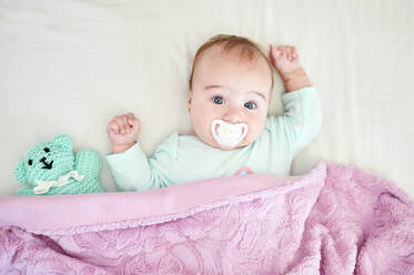 Portrait of baby girl with pacifier and cuddly toy lying on bed - KIJF03119