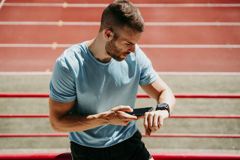 Male athlete in stadium checking smartwatch and smartphone - EBBF00233