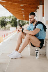 Male athlete with headphones, smartphone and smartwatch having a break sitting on grandstand in stadium - EBBF00235