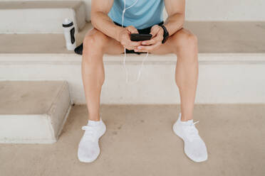 Male athlete having a break sitting on grandstand in stadium using smartphone - EBBF00236