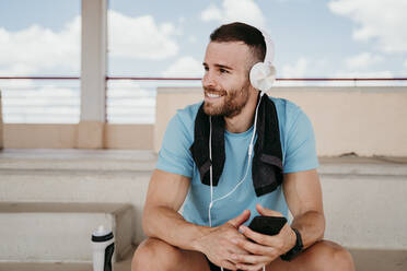 Male athlete with headphones and smartphone having a break sitting on grandstand in stadium - EBBF00237