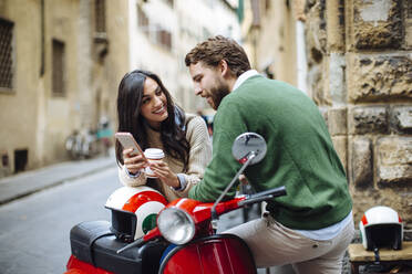 Lächelnde Frau zeigt ihrem Freund das Smartphone, während sie sich auf die Vespa lehnt - SODF00749