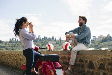 Woman photographing boyfriend sitting on retaining wall - SODF00758