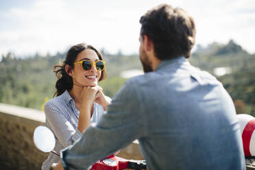 Lächelnde Frau mit Sonnenbrille im Gespräch mit ihrem Freund auf einer Vespa sitzend - SODF00759