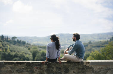 Couple looking at landscape while sitting on retaining wall against sky - SODF00763