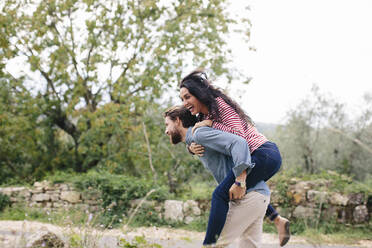 Happy man piggybacking girlfriend while running outdoors - SODF00778