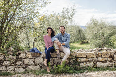 Smiling young couple sitting on retaining wall against trees - SODF00781