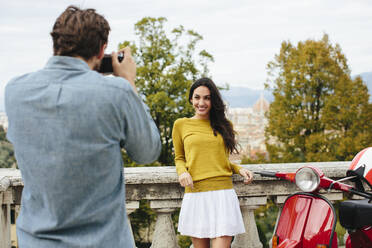 Young man photographing smiling girlfriend standing by railing - SODF00788