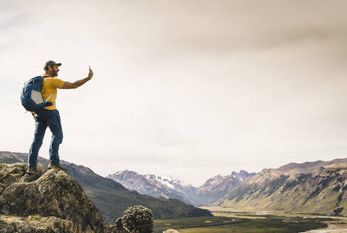 Älterer Mann, der ein Selfie mit seinem Smartphone macht, während er auf einem Felsen gegen den Himmel steht, Patagonien, Argentinien - UUF20679