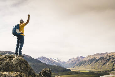 Mann mit erhobenem Arm, der auf einem Felsen stehend die Berge betrachtet, Patagonien, Argentinien - UUF20680