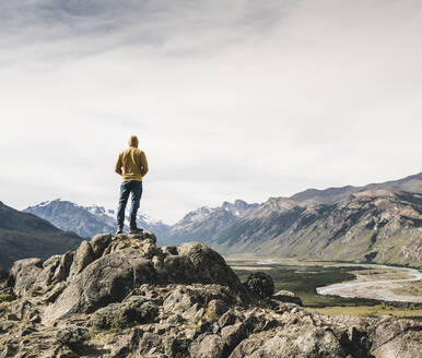 Älterer Mann mit Kapuze, der auf einem Felsen stehend die Berge gegen den Himmel betrachtet, Patagonien, Argentinien - UUF20681