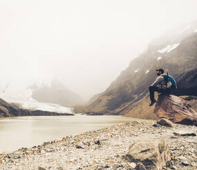 Älterer Mann mit Rucksack, der auf einem Felsen sitzend den See betrachtet, Patagonien, Argentinien - UUF20696