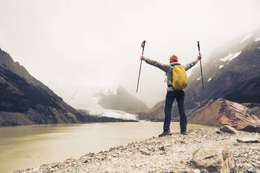 Man with arms outstretched holding hiking poles standing by lake at Patagonia, Argentina - UUF20701