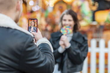 Mann fotografiert Freundin beim Essen eines Lutschers mit dem Smartphone im Vergnügungspark - WPEF03097