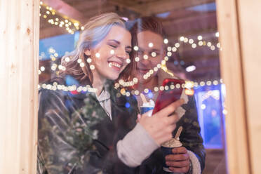 Junges Paar benutzt Smartphone in einem Geschäft im Vergnügungspark, gesehen durch ein Fenster - WPEF03106