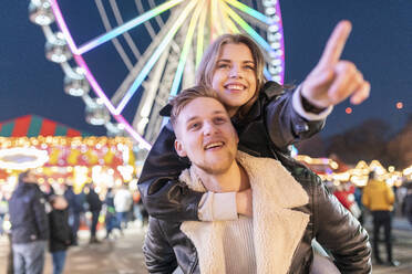 Man piggybacking girlfriend pointing while standing in illuminated amusement park at night - WPEF03116