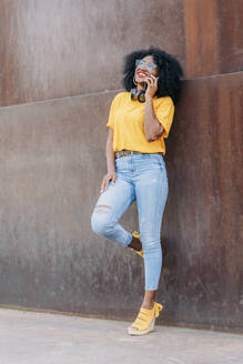 Smiling woman with afro hair and glasses using smartphone leaning on wall - JAF00003