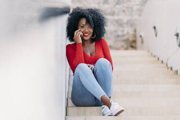 Smiling woman with afro hair using smartphone sitting on stairs - JAF00005