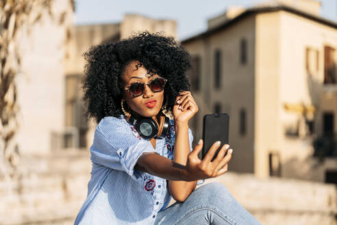 Smiling woman with afro hair and sunglasses taking a selfie with her smartphone - JAF00010