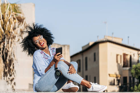 Lachende Frau mit Afro-Haar und Brille, die ein Smartphone benutzt - JAF00012
