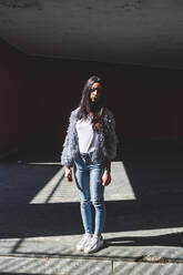 Junge Frau mit Jacke, die an einem sonnigen Tag vor einem Gebäude steht - MEUF01022