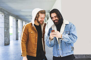 Junger Mann zeigt einem männlichen Freund sein Smartphone, während er an der Wand steht - MEUF01026
