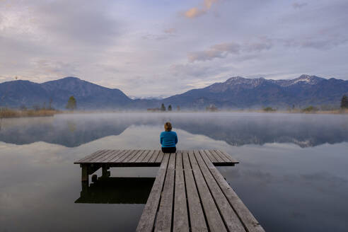 Deutschland, Bayern, Schlehdorf, Frau sitzt am Ende des Stegs und bewundert den Eichsee bei nebligem Morgengrauen - LBF03091