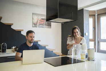 Mature man working over laptop while smiling woman preparing drink on kitchen island - MTBF00448