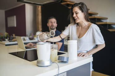 Man working over laptop talking with woman preparing drink on kitchen island - MTBF00449