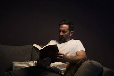 Mature man reading book while sitting on sofa against wall at home - MTBF00459